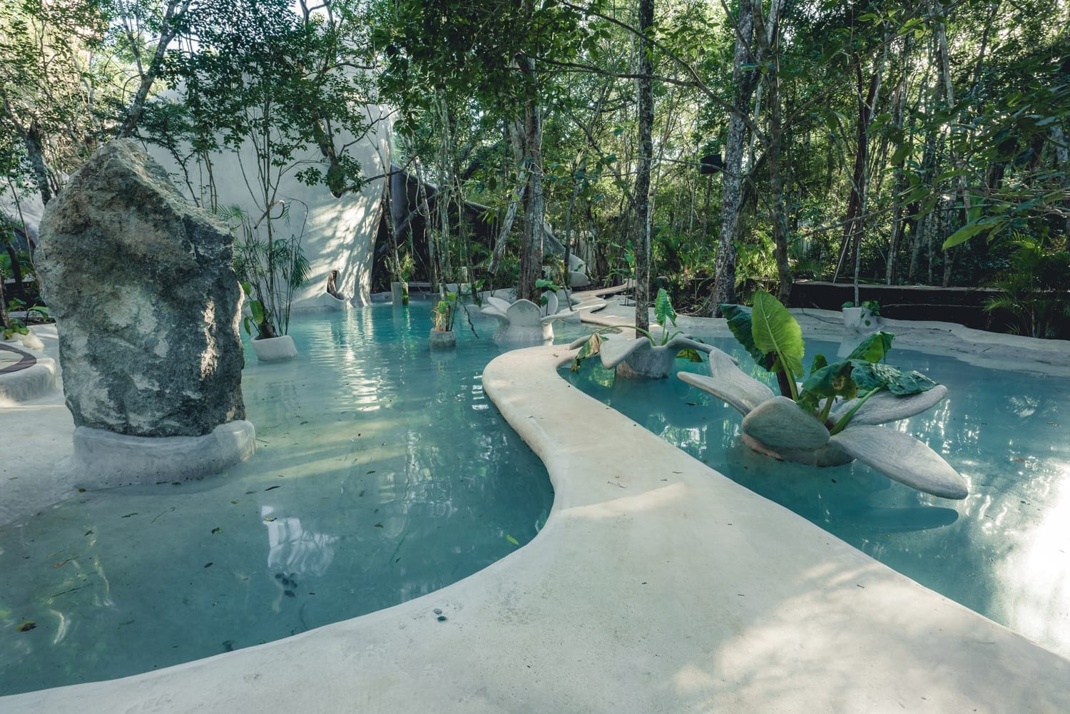 Serene pool surrounded by lush trees and plants, featuring stone pathways and a large boulder, creating a tranquil oasis.