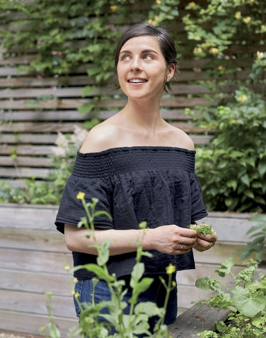 Person in an off-shoulder black top standing in a garden, holding plants with greenery in the background.