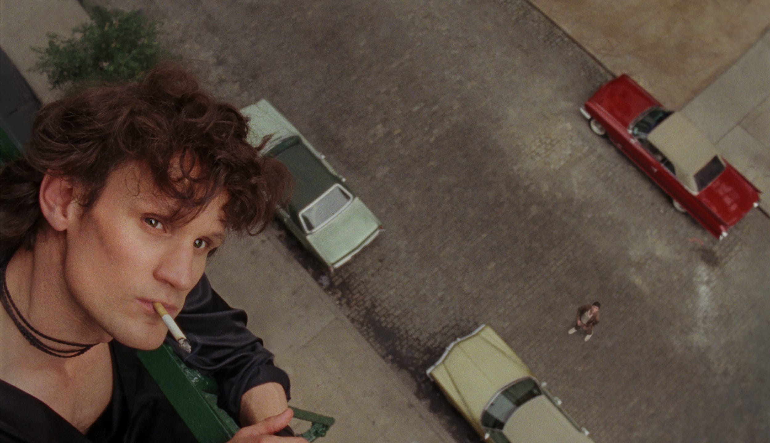 Person leaning out of window smoking, looking down at street with vintage cars parked below.