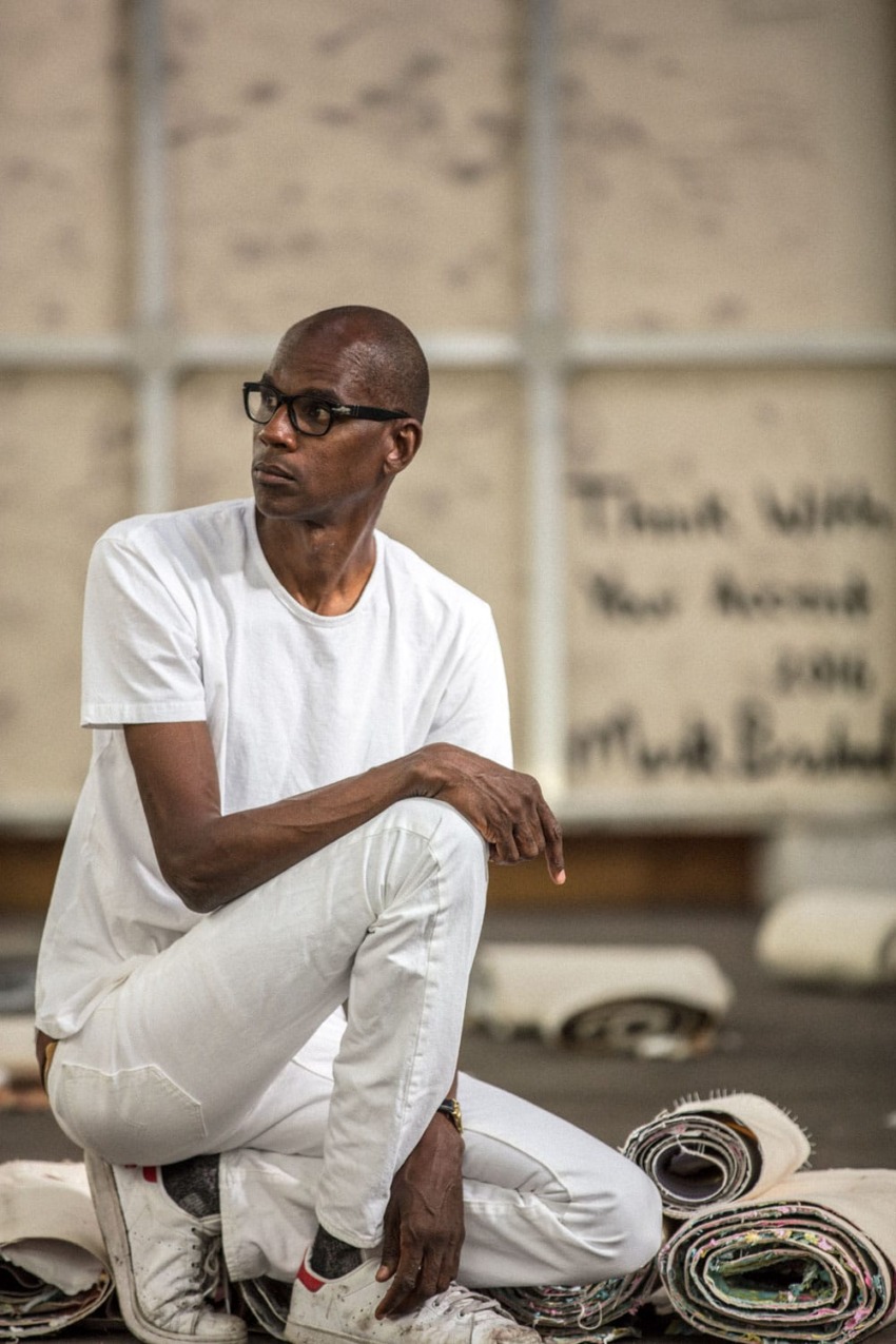 Person in white outfit sitting on rolled canvas in an art studio, thoughtful expression, glasses, varied background textures