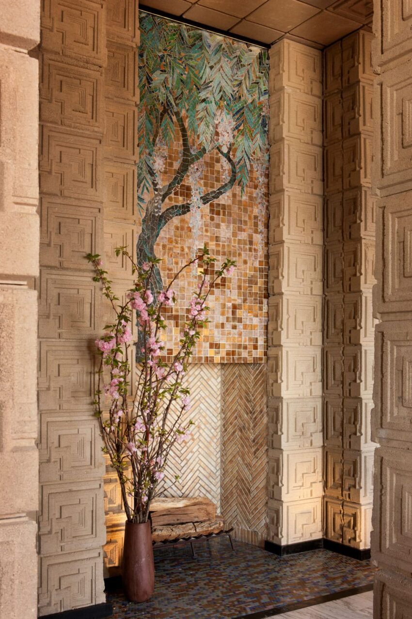 Intricate mosaic mural with tree design above a vase of pink blossoms, surrounded by textured patterned walls.
