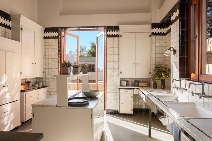Spacious kitchen with white tiles, open wooden doors, counters, plants, and natural sunlight streaming in.