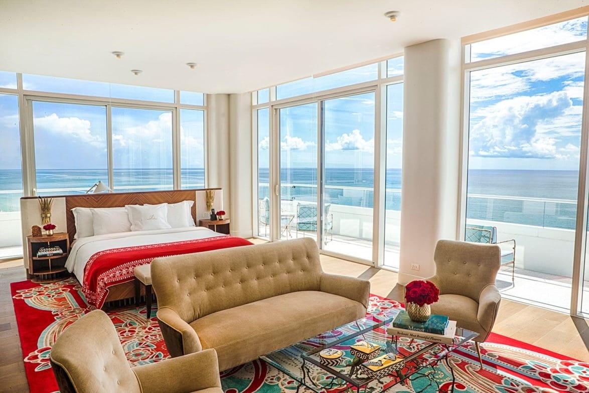 Luxurious ocean-view bedroom with large windows, modern furniture, vibrant decor, and a sunlit balcony.