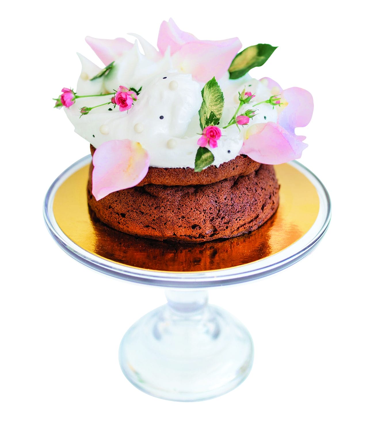Chocolate cake on a glass stand topped with white frosting, pink petals, and small pink flowers.