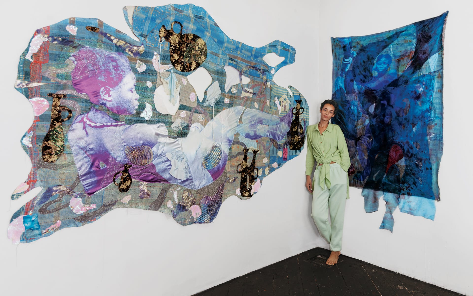 Artist in green outfit stands beside vibrant, abstract wall art with blue and pink hues, featuring intricate patterns and textures.