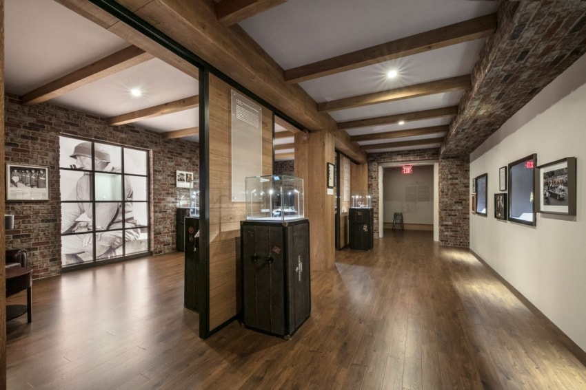 Museum hallway with glass display cases, wooden beams, and framed photographs on brick and white walls.
