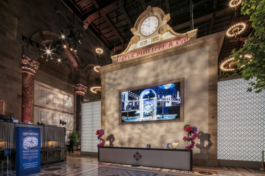 Elegant interior with clock and large screen displaying an intricate timepiece, surrounded by floral arrangements and ambient lighting.