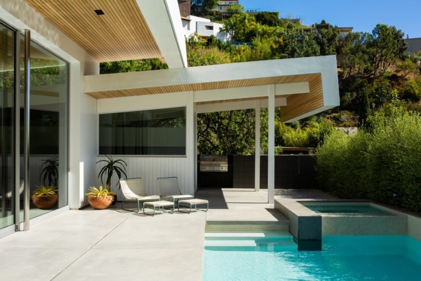 Modern house with a pool, patio chairs, and lush greenery in a sunny outdoor setting.