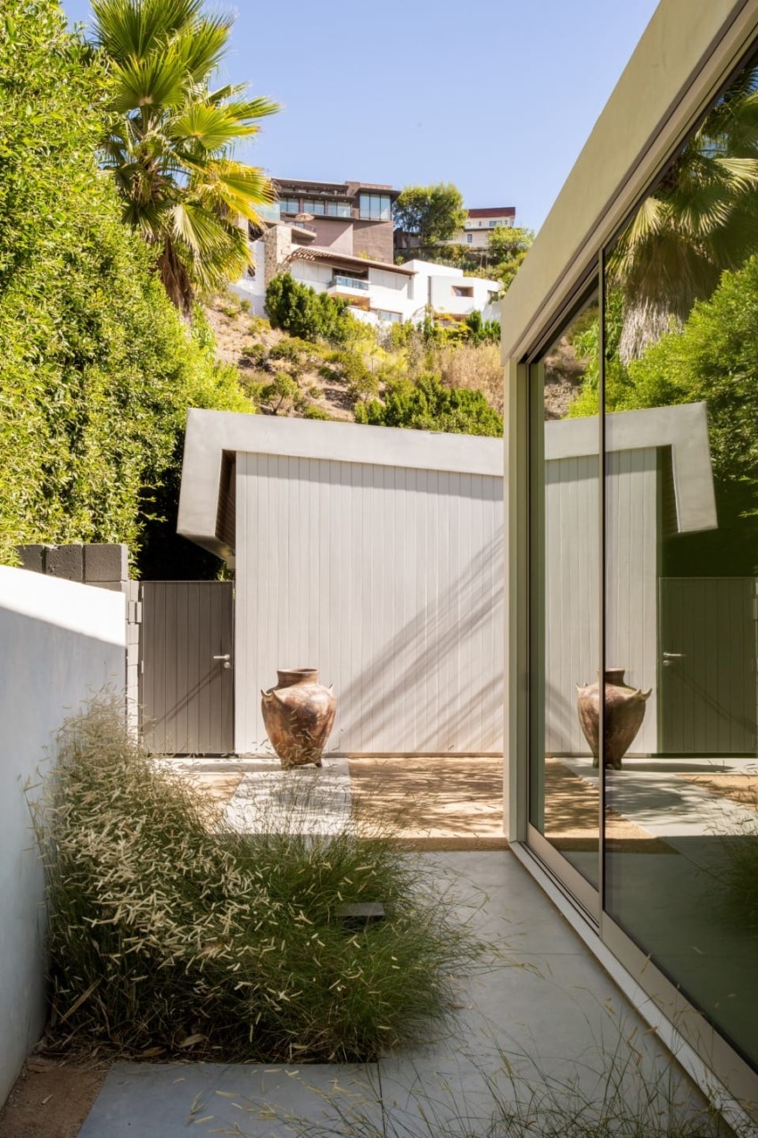 modern house exterior with large patio doors, potted plants, and landscaped greenery, hillside homes visible in background