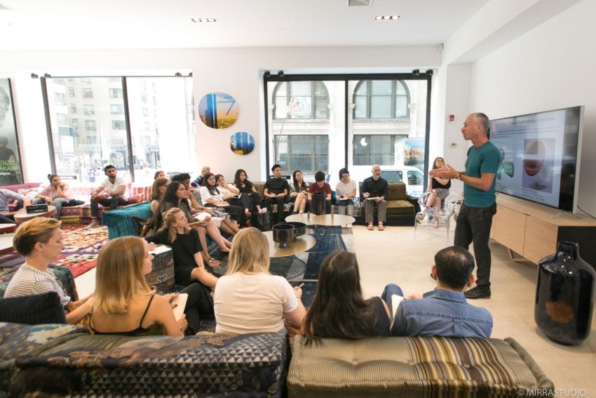 A man giving a presentation to a group of seated people in a modern, well-lit room with large windows.