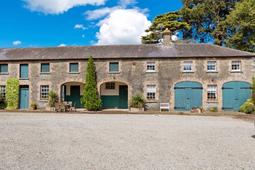 Historic stone building with blue shutters and arched entryways, surrounded by trees and a gravel courtyard under a blue sky.