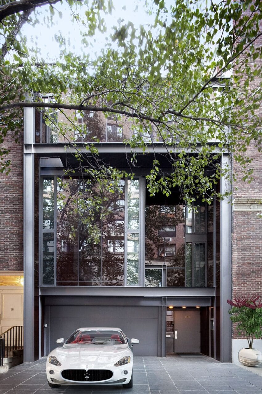 White Maserati parked in front of modern glass garage with brick exterior and tree branches above.
