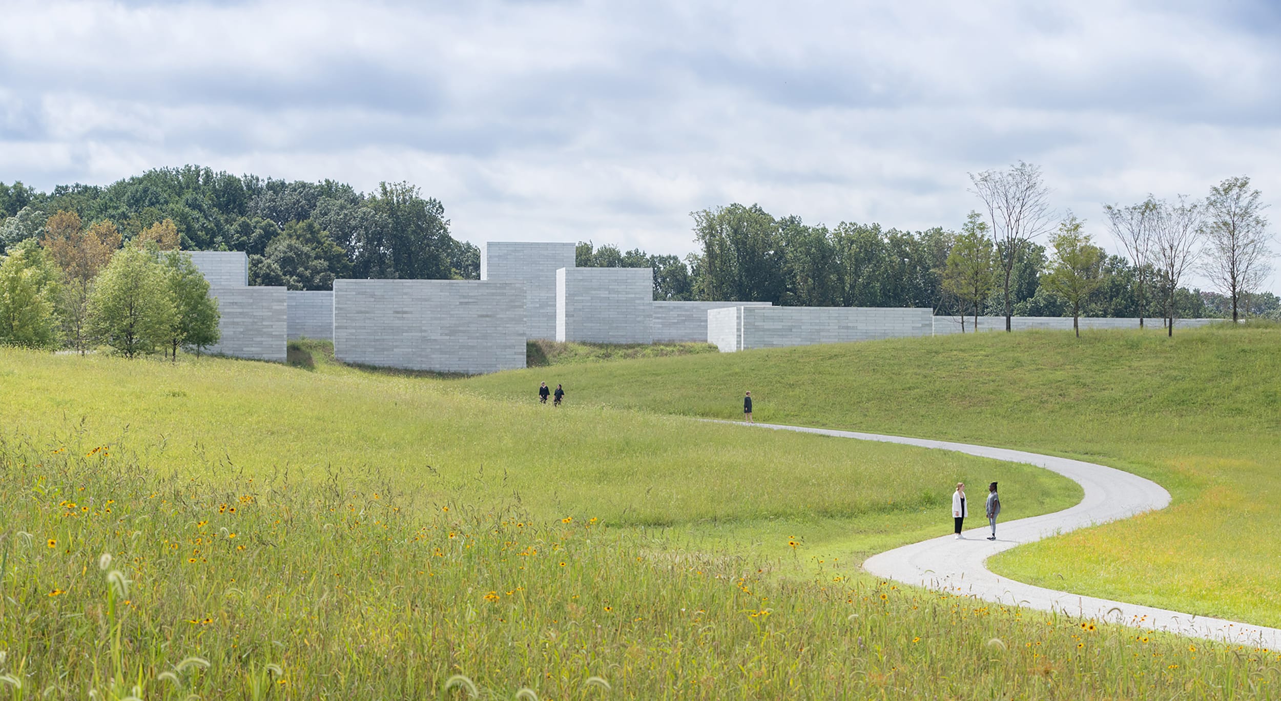 Pathway through a grassy field leading to modern white buildings surrounded by trees under a cloudy sky