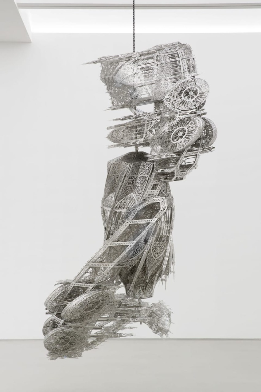 Intricate metallic sculpture hanging in a gallery, showcasing complex patterns and details in a twisting vertical form.