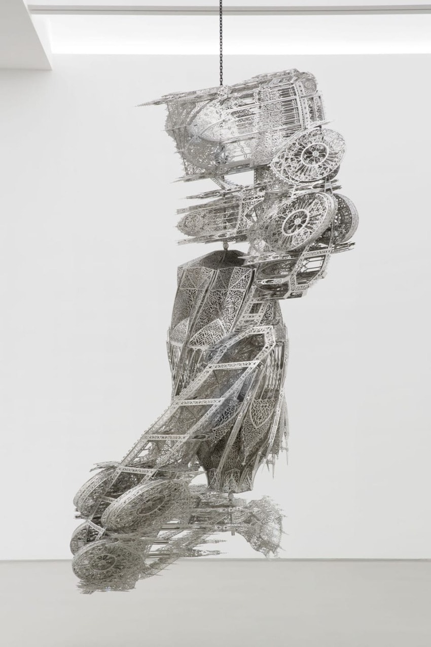 Intricate metallic sculpture hanging in a gallery, showcasing complex patterns and details in a twisting vertical form.