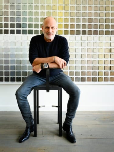 Man sitting on a chair in front of a wall covered with colored swatches, wearing a black sweater and jeans, arms crossed.