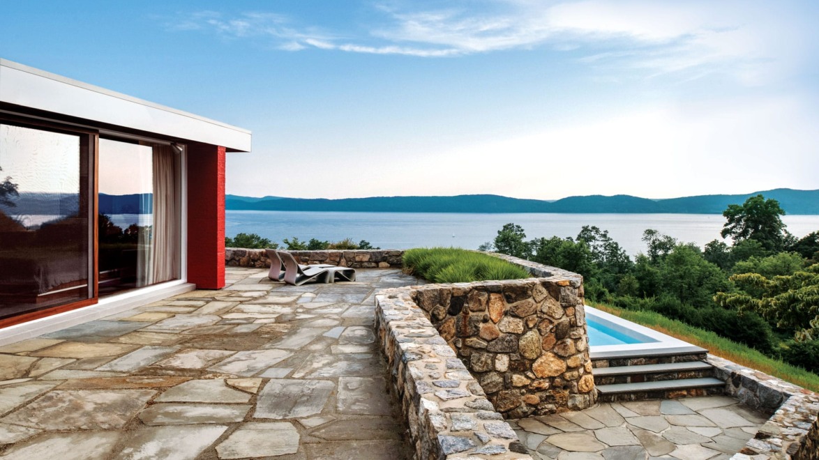 Modern patio with stone wall overlooking a serene lake and green hills under a blue sky.