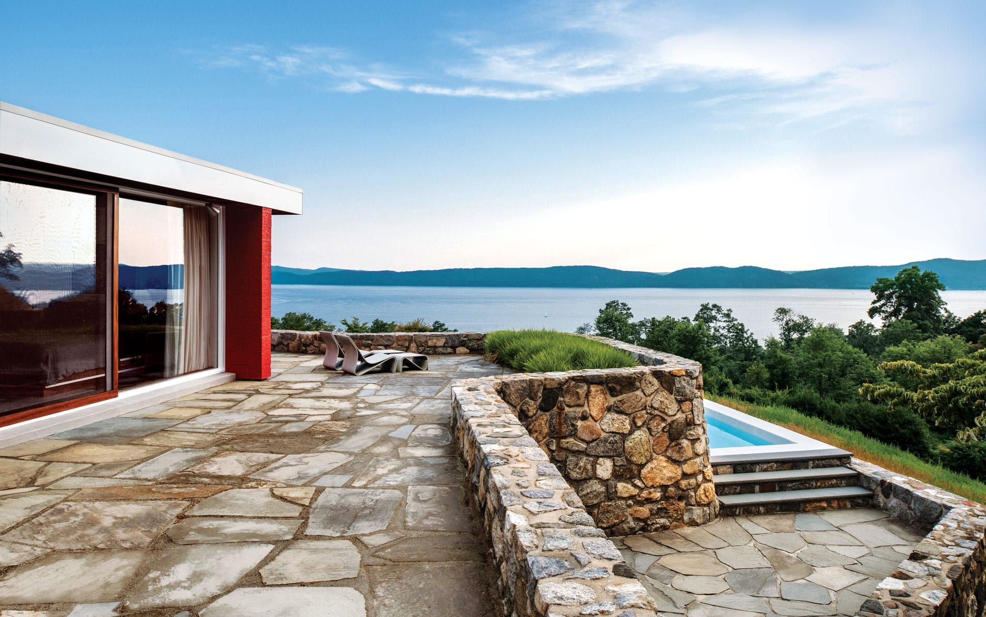 Modern patio with stone wall overlooking a serene lake and green hills under a blue sky.