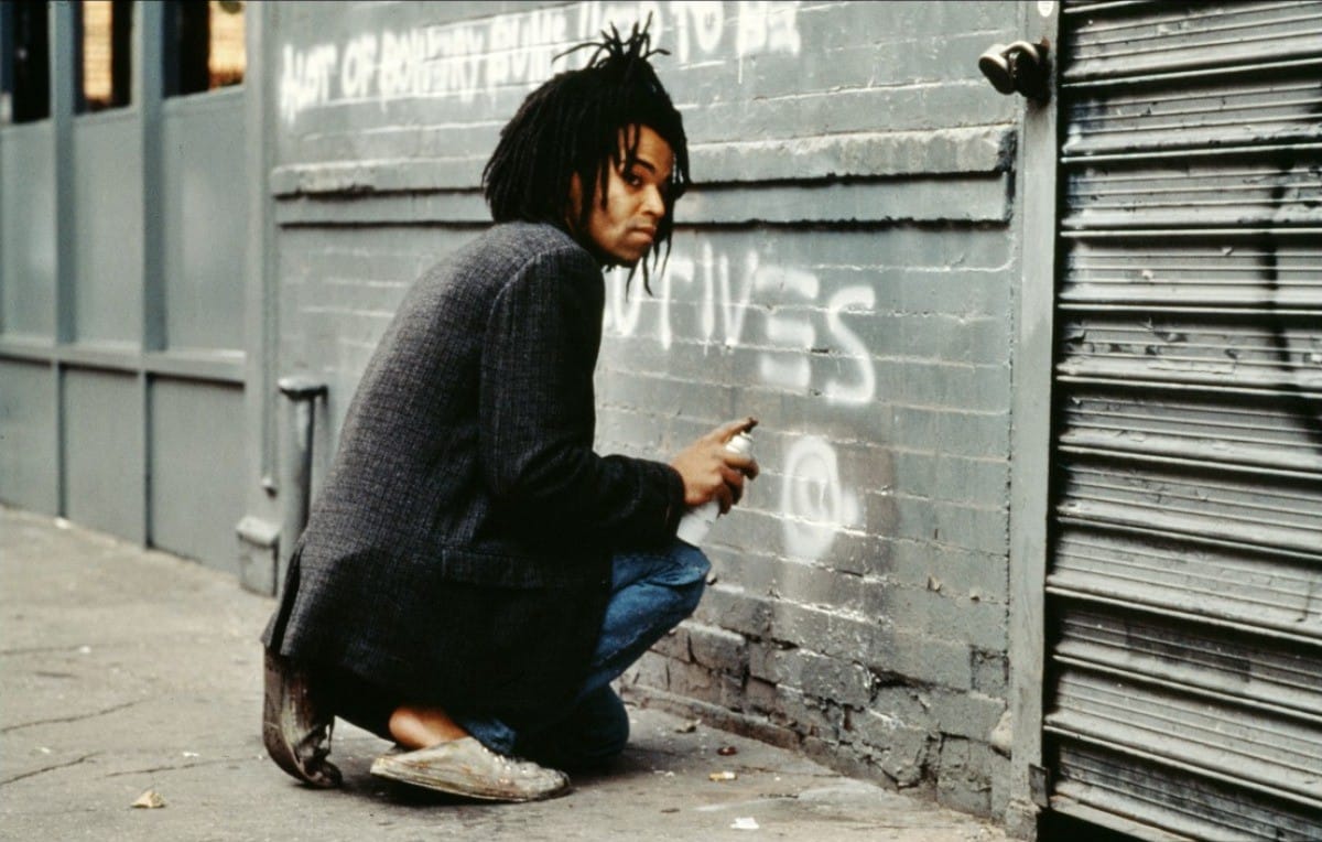 Person crouching, spray painting graffiti on a wall in an urban setting, wearing casual clothing and sneakers.