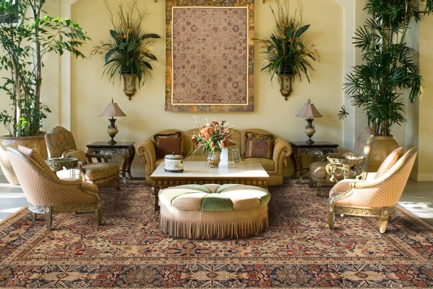 Elegant living room with patterned rug, cushioned chairs, sofa, coffee table, wall tapestry, and decorative plants.