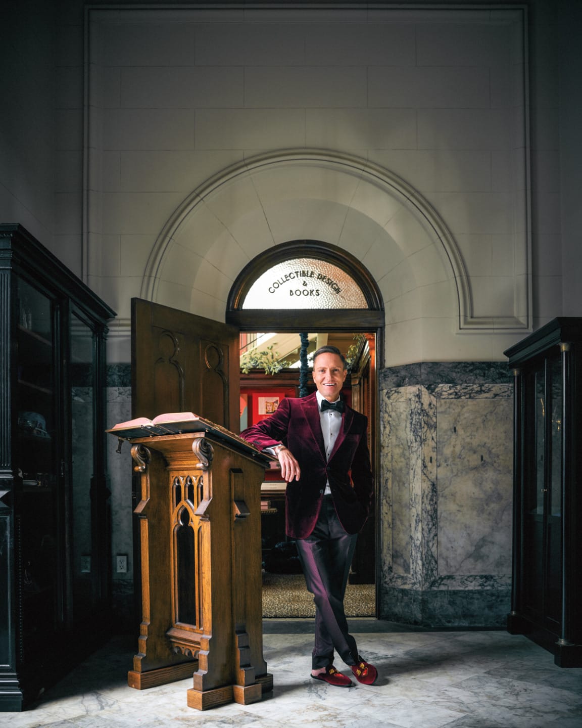 Man in a velvet suit leans against a wooden lectern in a luxurious room, surrounded by books, marble, and an arched doorway.