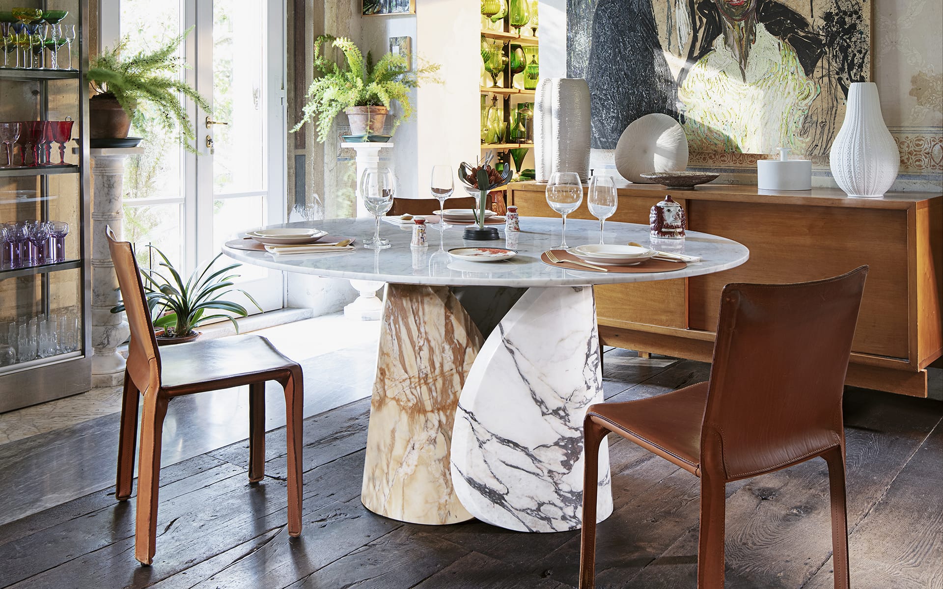 Round marble dining table set with glassware, surrounded by leather chairs in a modern, plant-filled room.