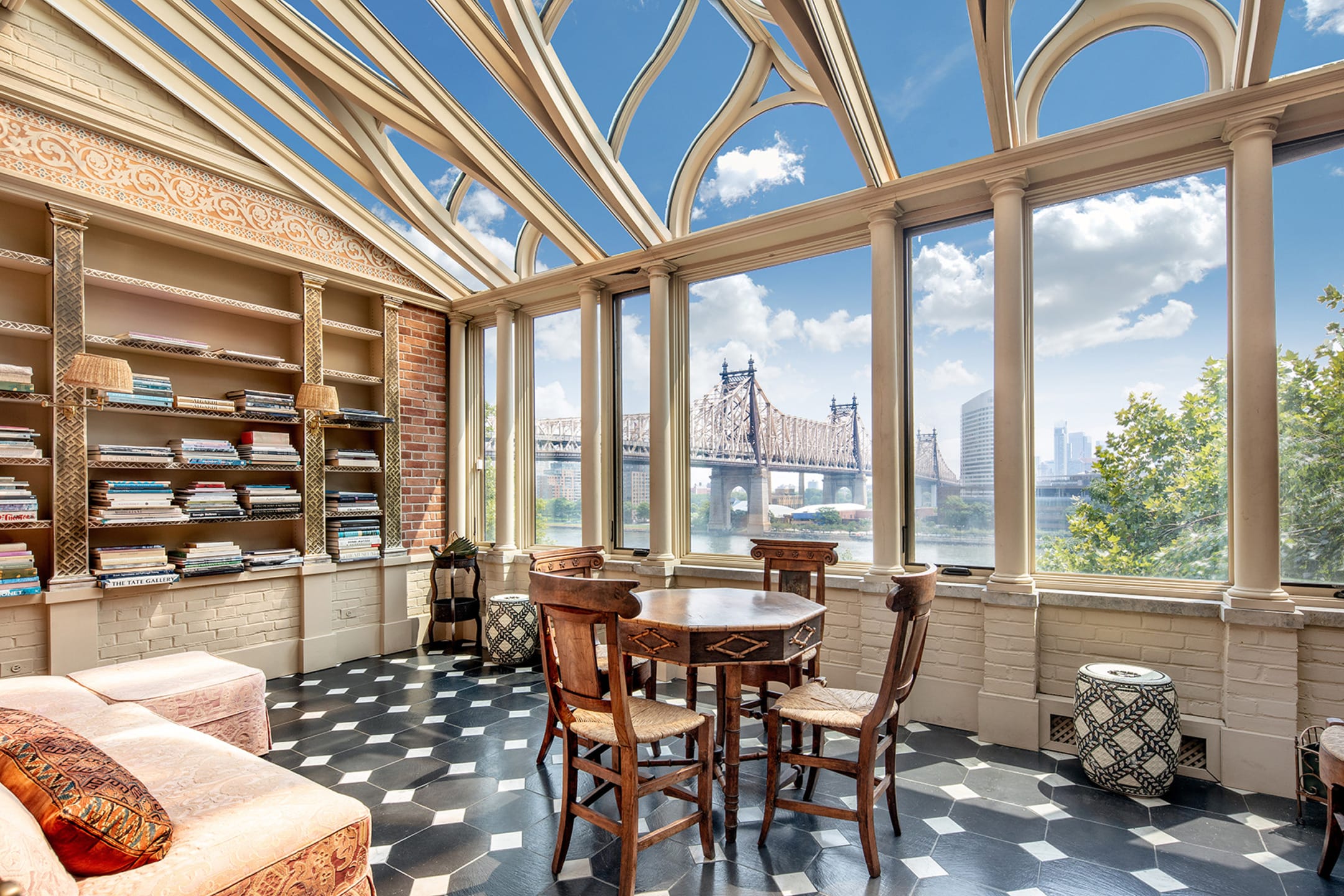 Sunlit room with a round table, wooden chairs, bookshelves, and large windows offering a view of a bridge and blue sky.