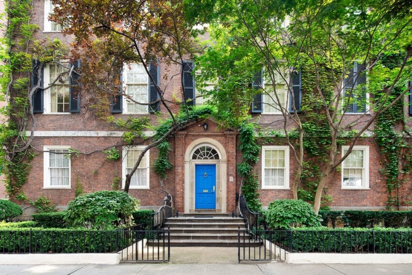 Historic brick townhouse with ivy, blue door, black shutters, and lush green trees and shrubs lining the front pathway.