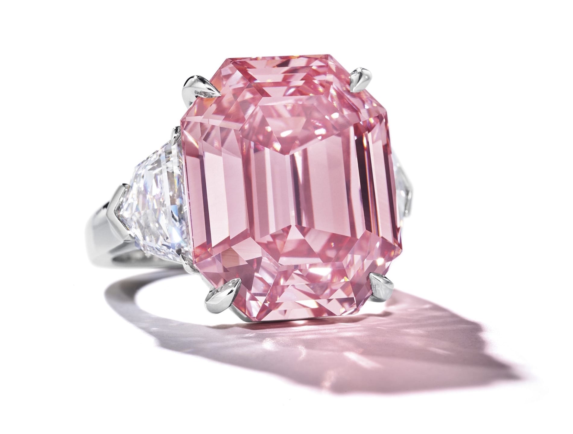 A large pink diamond ring with an emerald cut and white diamond side stones on a reflective surface.