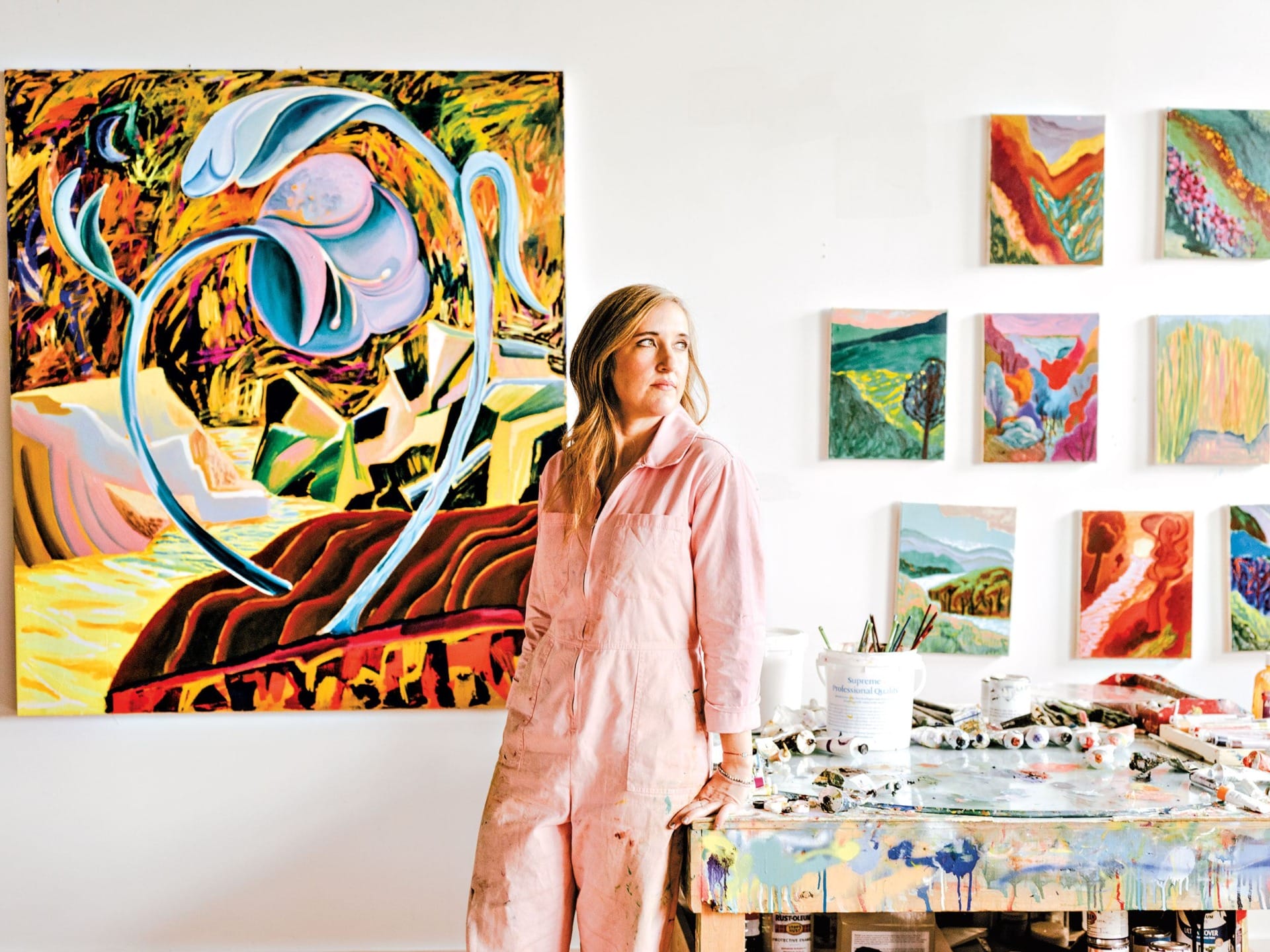 Artist in studio wearing pink overalls, standing next to colorful abstract paintings on wall and messy table with art supplies.