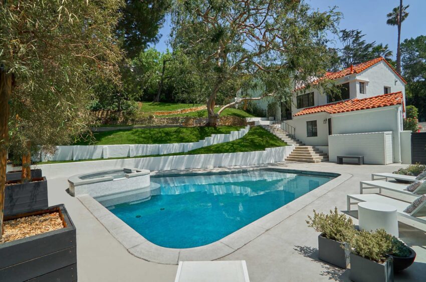 Backyard view of a house with a pool, hot tub, green lawn, trees, and a modern white house with a red-tiled roof.