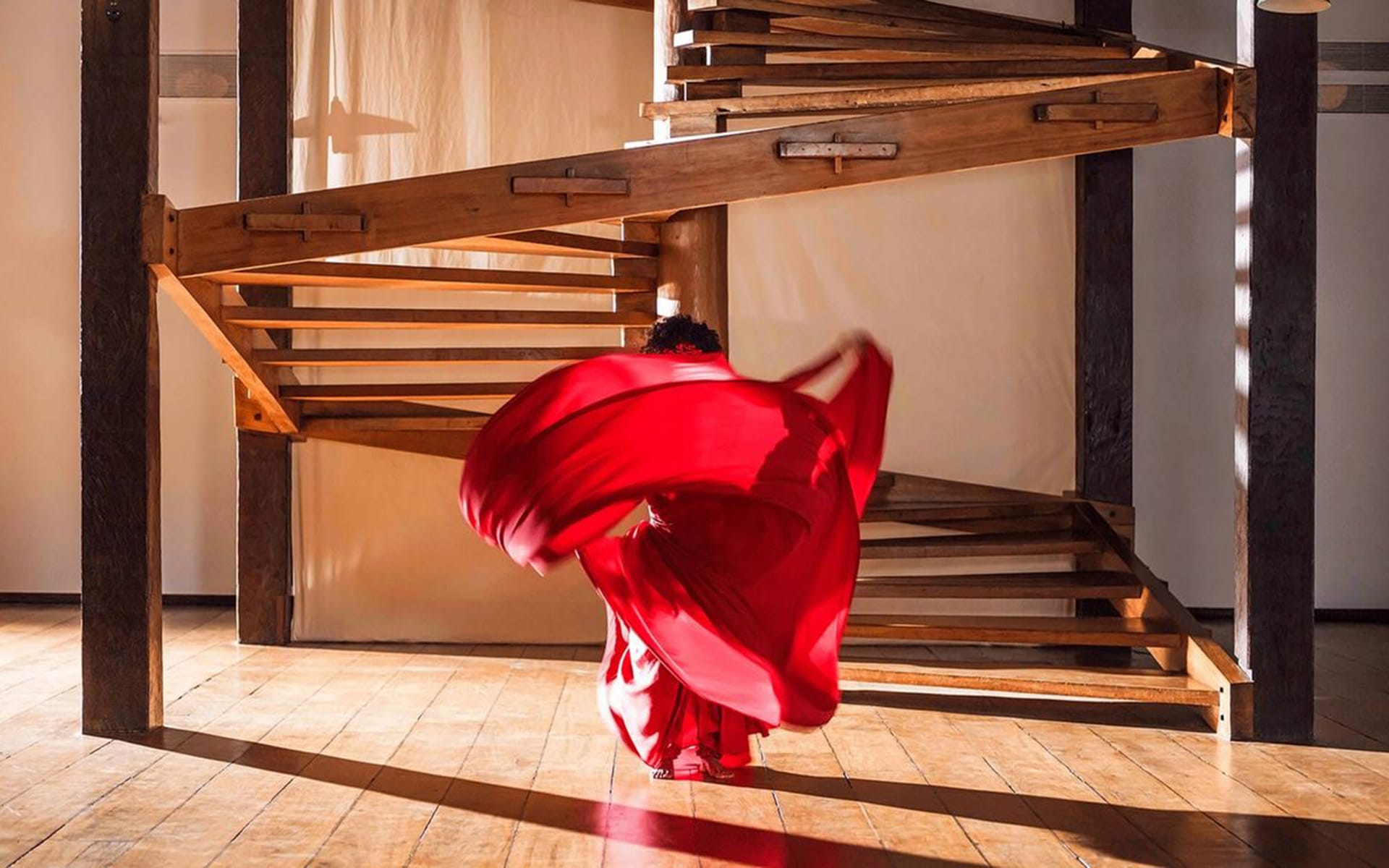 Person dancing in vibrant red dress on wooden floor with spiral staircase background, creating a dynamic flowing motion.