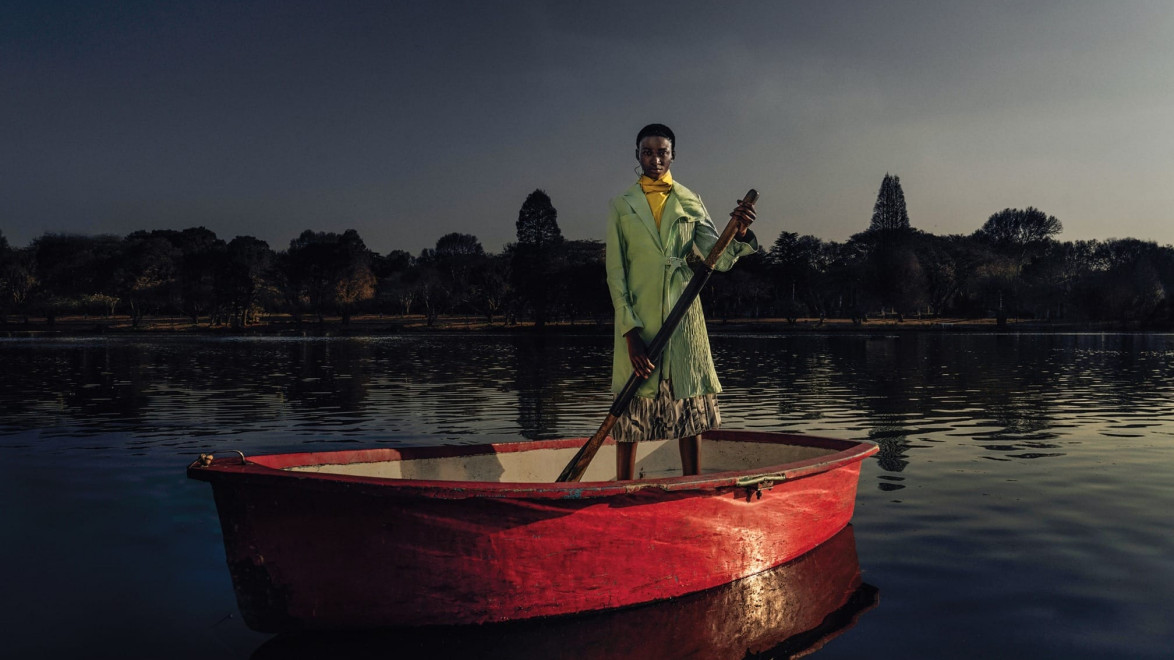 Person in a green coat standing in a red rowboat on a calm lake with trees in the background at dusk.