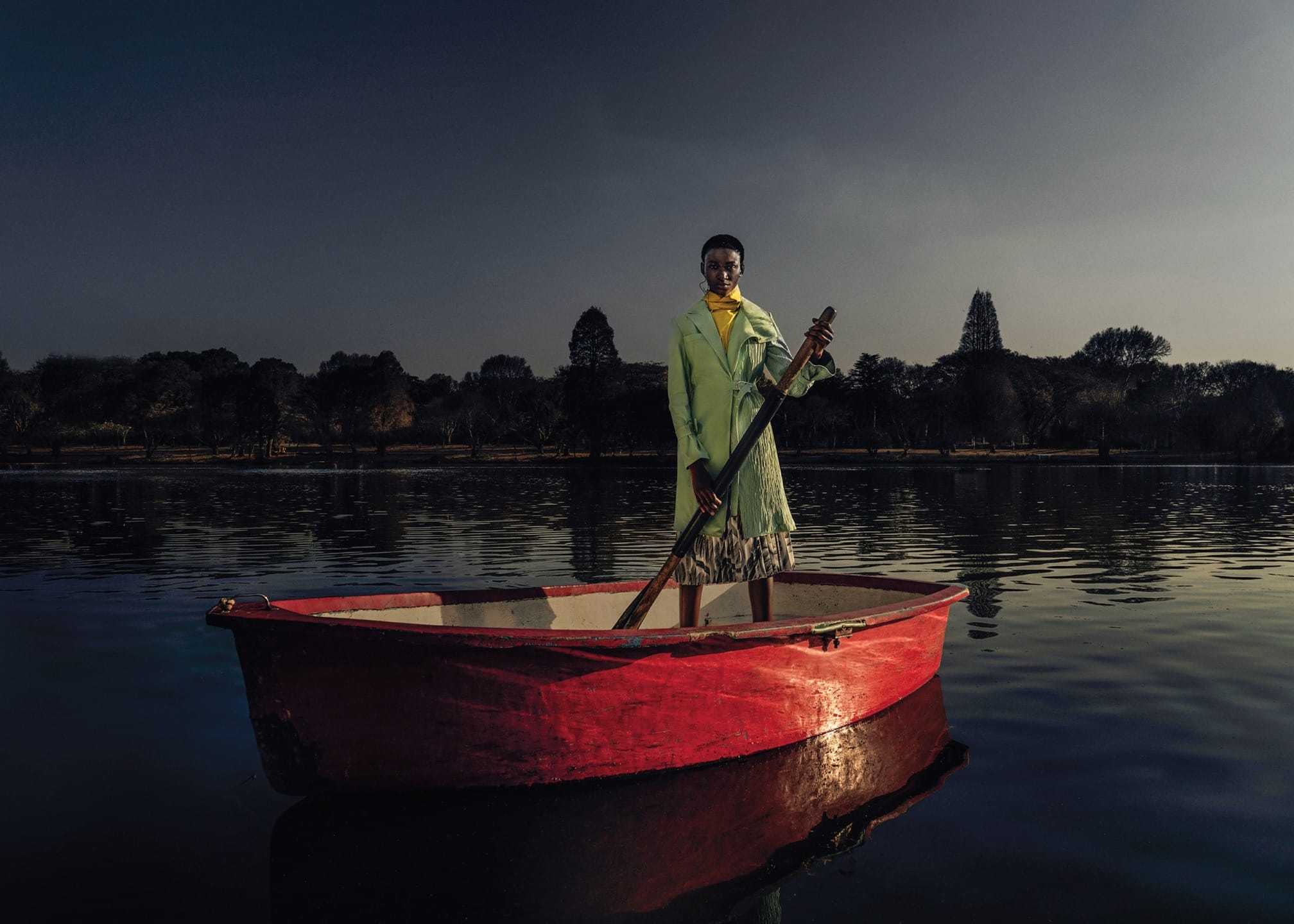 Person in a green coat standing in a red rowboat on a calm lake with trees in the background at dusk.