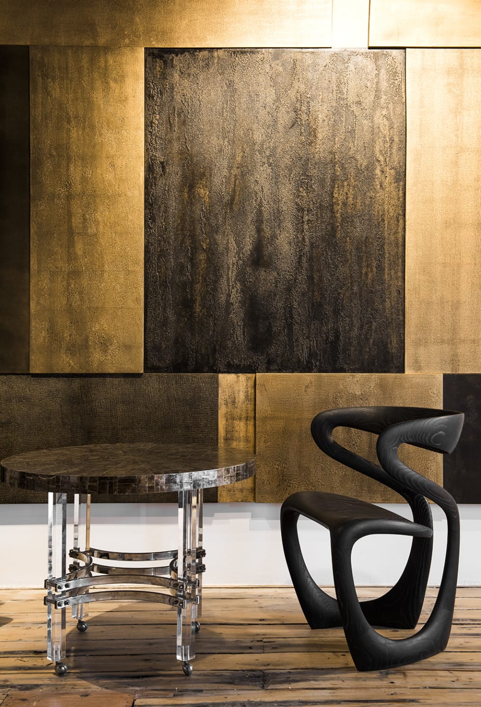 Modern art gallery with textured gold panels, clear table, and abstract black chair on wooden floor.