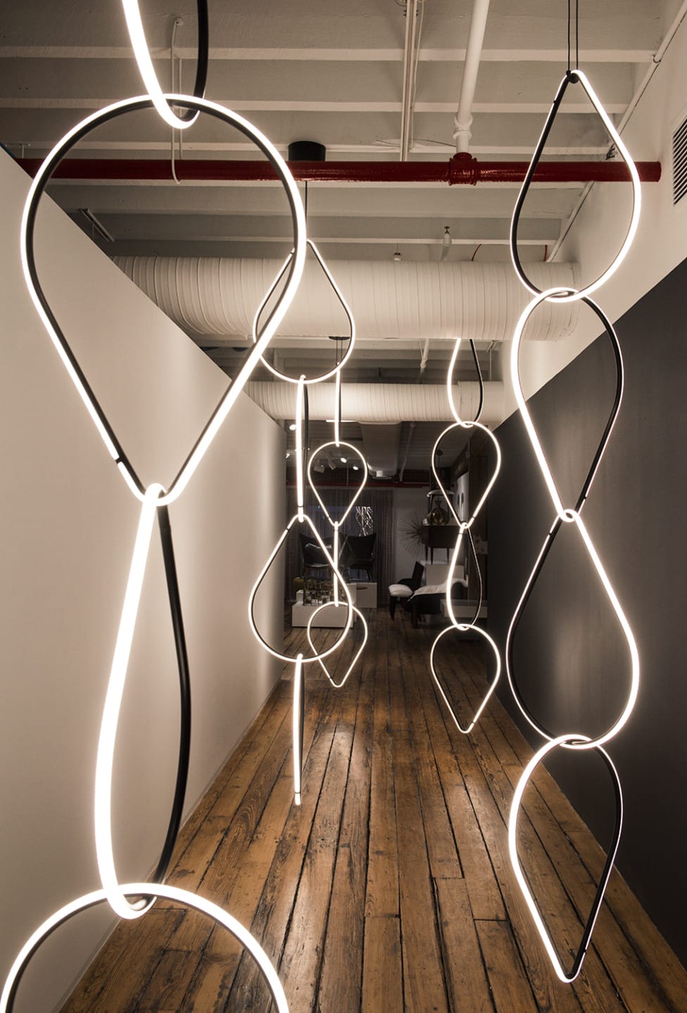 Modern art installation with illuminated loops hanging in a dimly lit hallway with wooden floors and industrial ceiling.