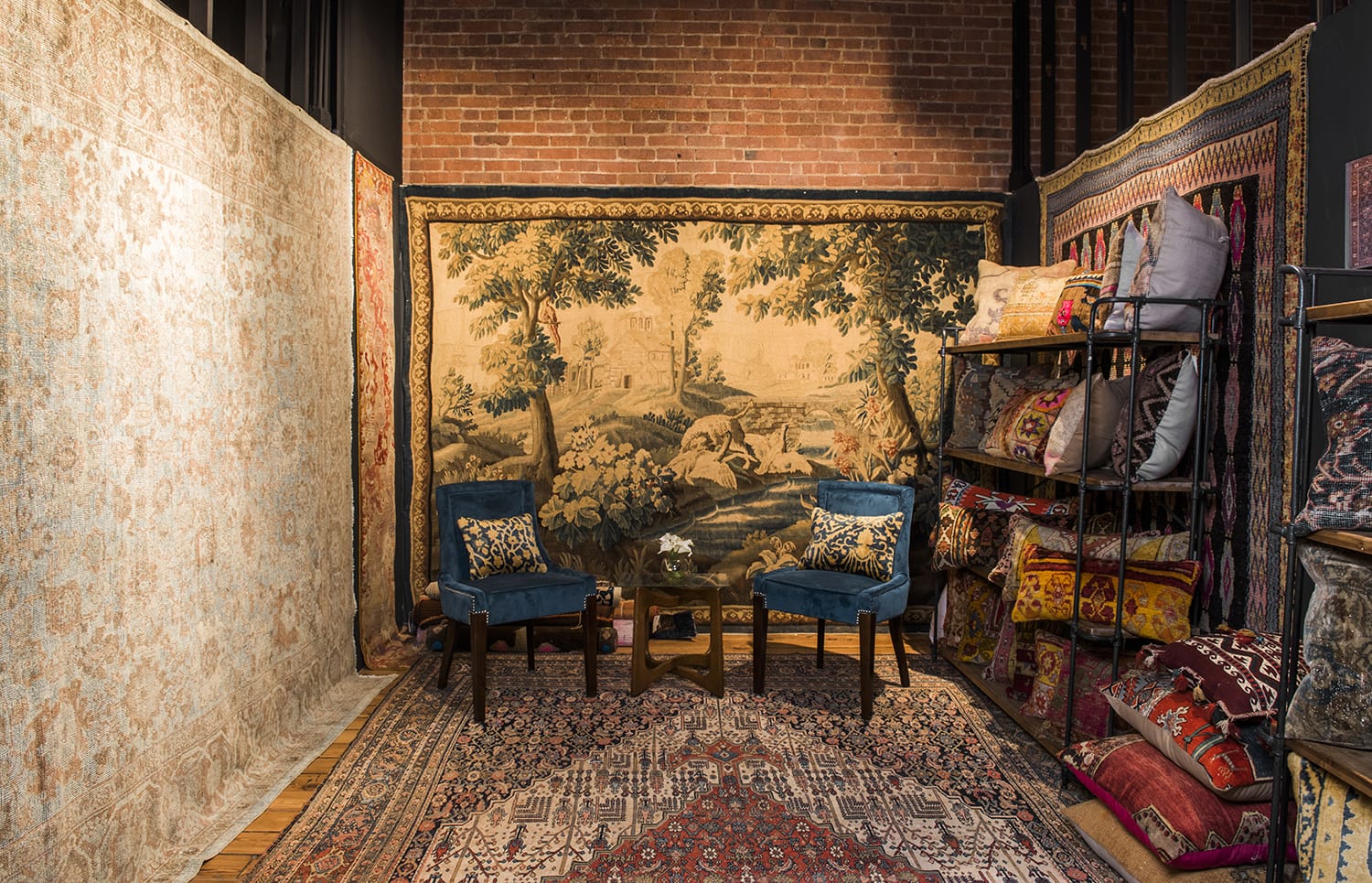Cozy room with decorative tapestry, vintage rugs, blue chairs, and colorful cushions on shelves against a brick wall.