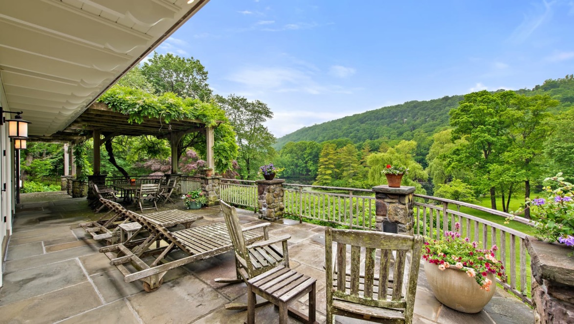 Spacious outdoor patio with wooden chairs, lush greenery, flowering plants, and distant hills under a clear blue sky.