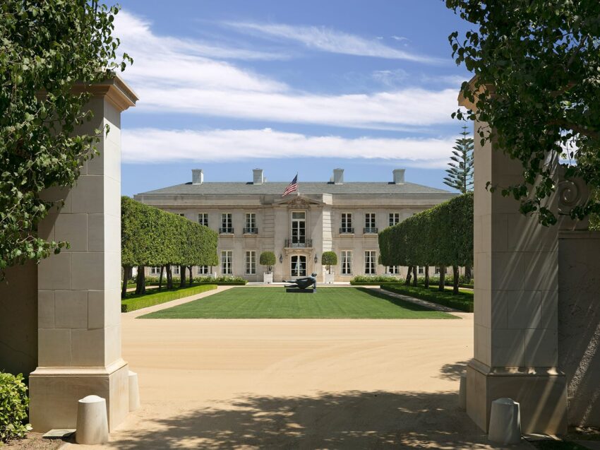 Elegant mansion with manicured lawn and trees, viewed through stone gate pillars under a clear blue sky.