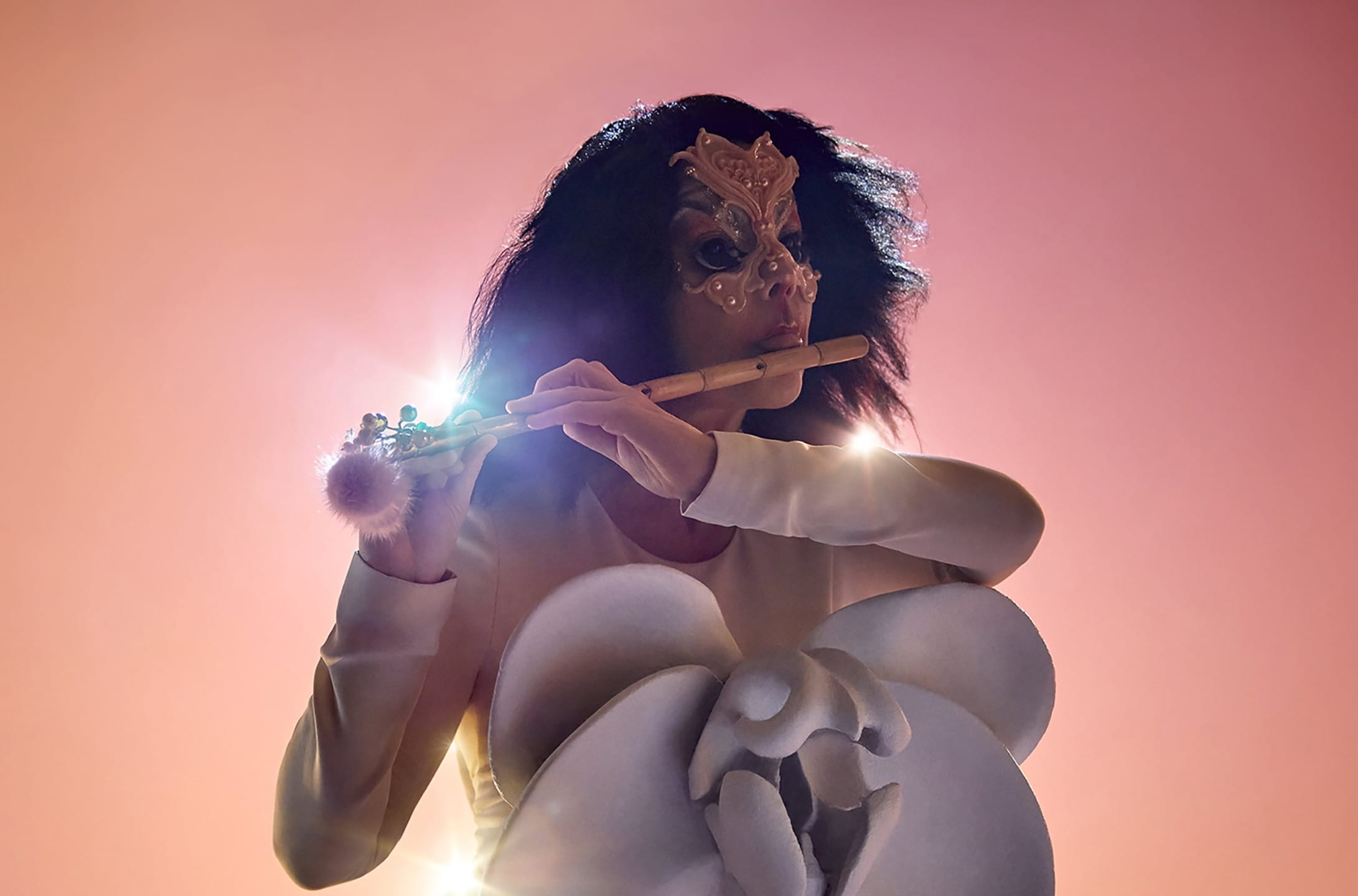 A person in a decorative mask plays a wooden flute against a pink backdrop, with light flaring in the background.