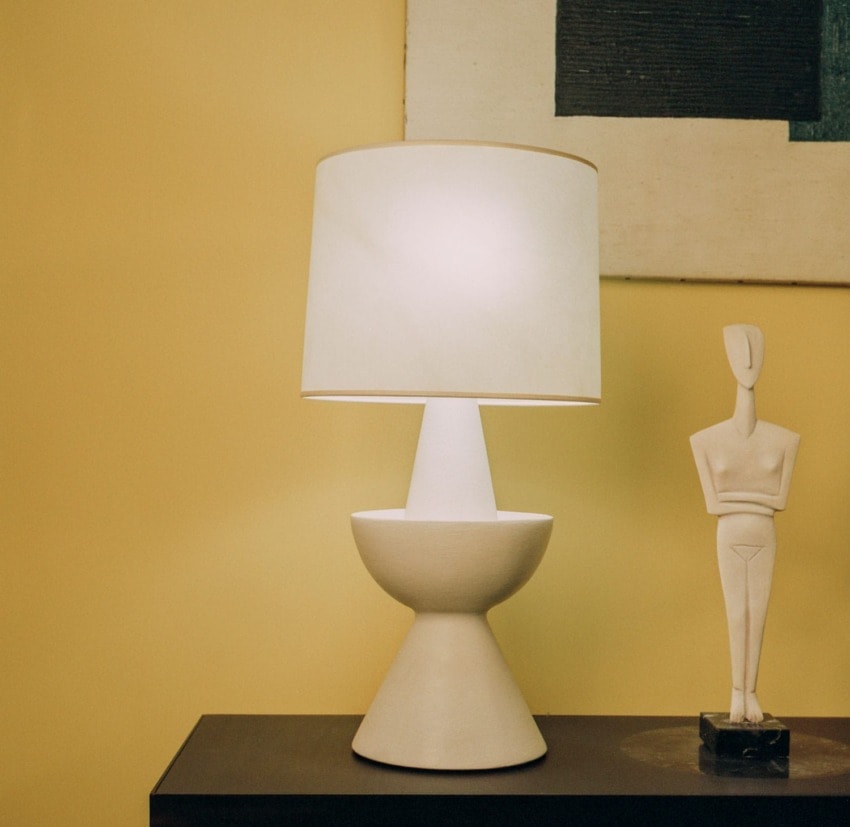 Modern white table lamp with a geometric base on a dark wooden surface, beside an abstract sculpture, against a yellow wall.