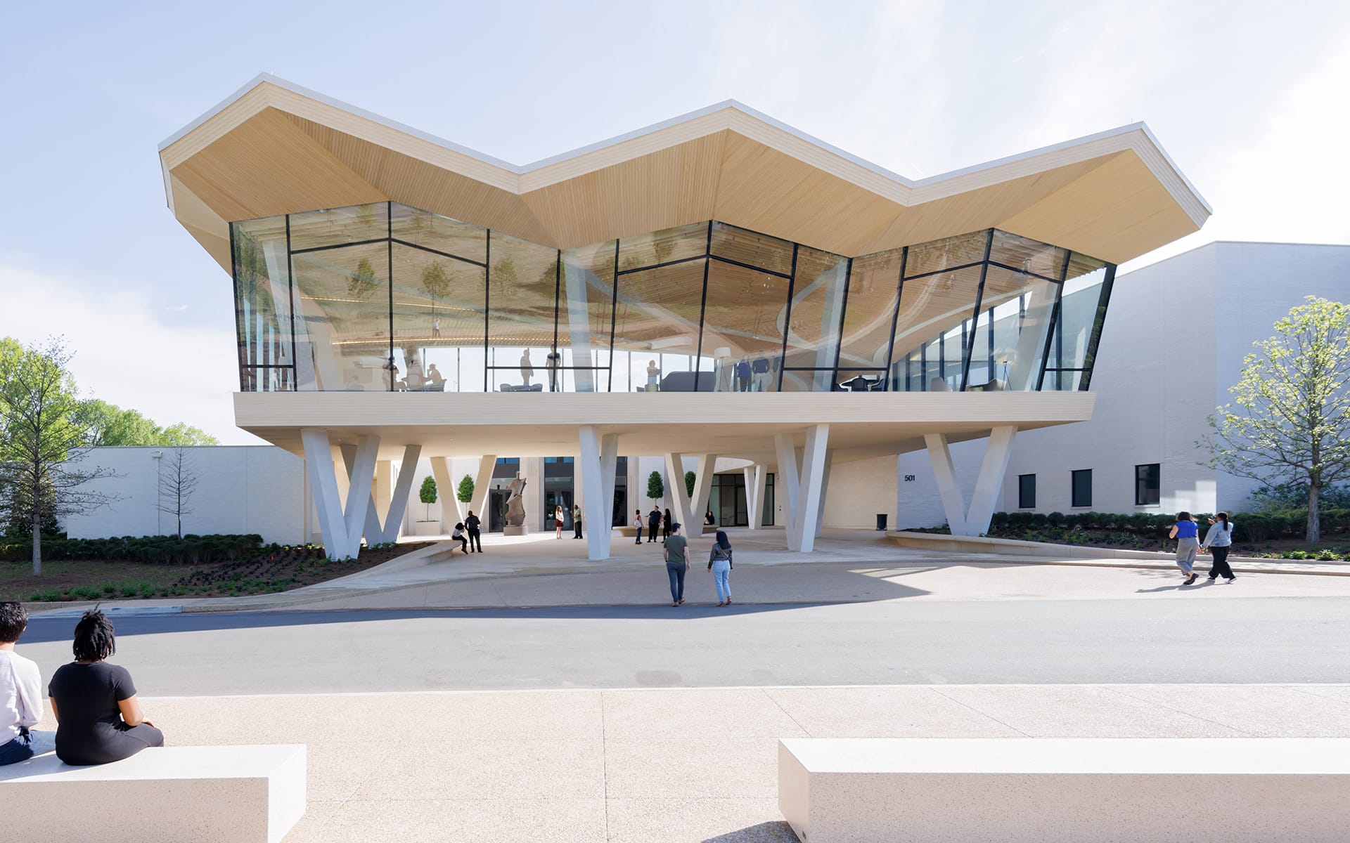 Modern building with unique roof design, glass facade, and people walking nearby, surrounded by trees and clear skies.