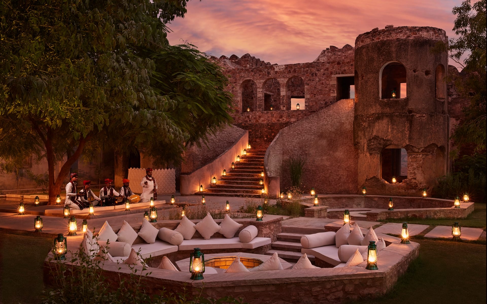 Outdoor seating area with cushions and lanterns at an ancient fort during sunset, musicians playing traditional instruments nearby.