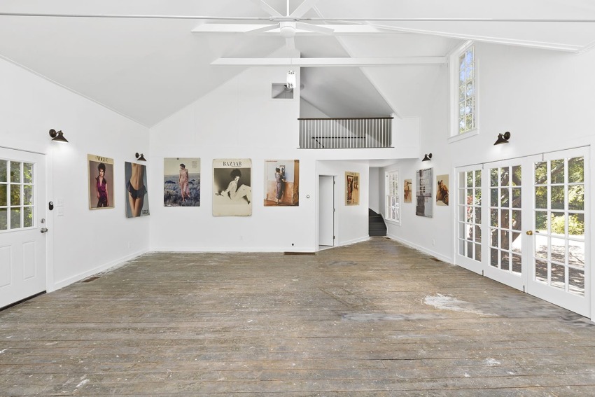 Spacious white art gallery interior with large windows, wooden floor, and various artworks displayed on the walls.