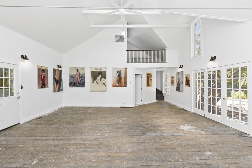 Spacious white art gallery interior with large windows, wooden floor, and various artworks displayed on the walls.