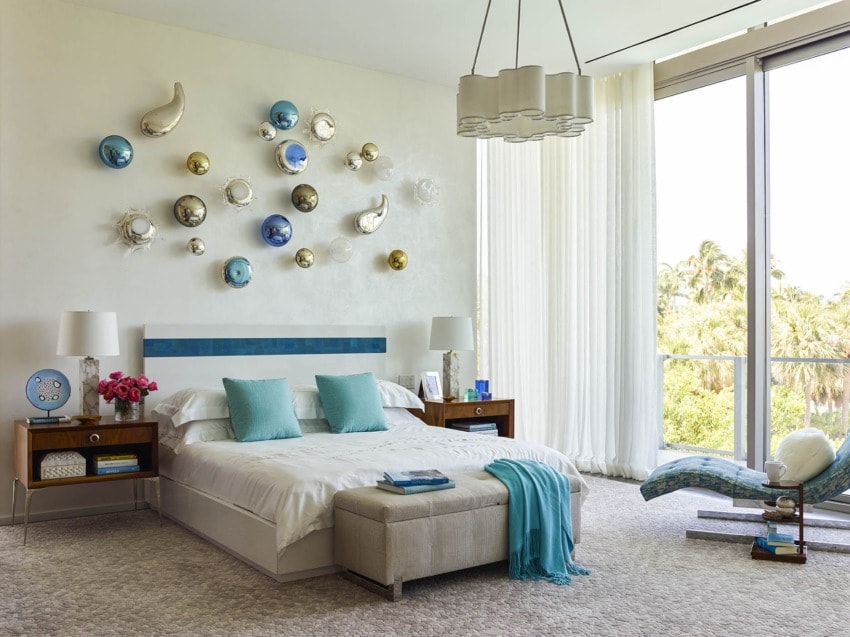 Modern bedroom with bright decor, large window, unique wall art, blue accents, and a comfortable bed with decorative pillows.