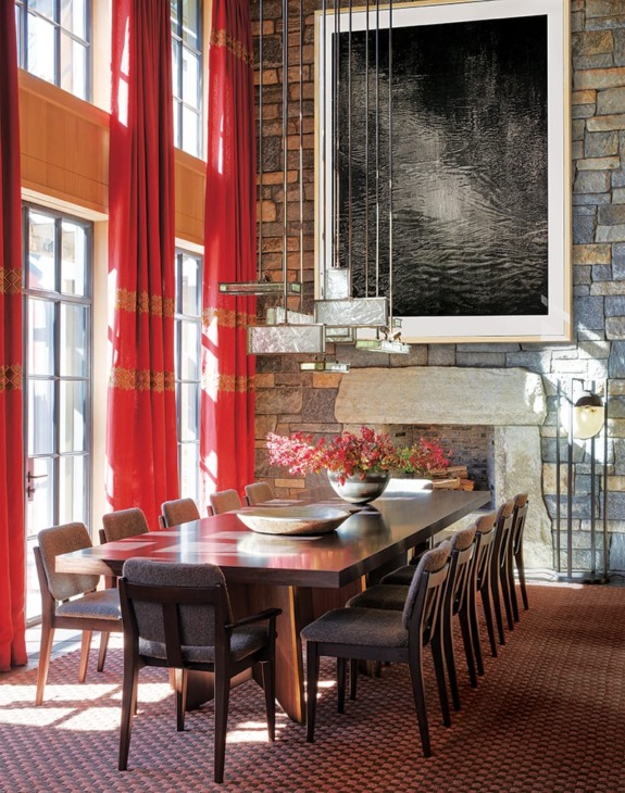 Spacious dining room with a wooden table, chairs, red curtains, stone walls, and contemporary art above a fireplace.