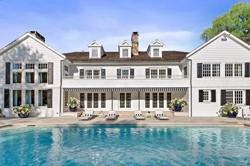 Luxurious white country house with large pool, spacious patio, and elegant landscaping under a clear blue sky.