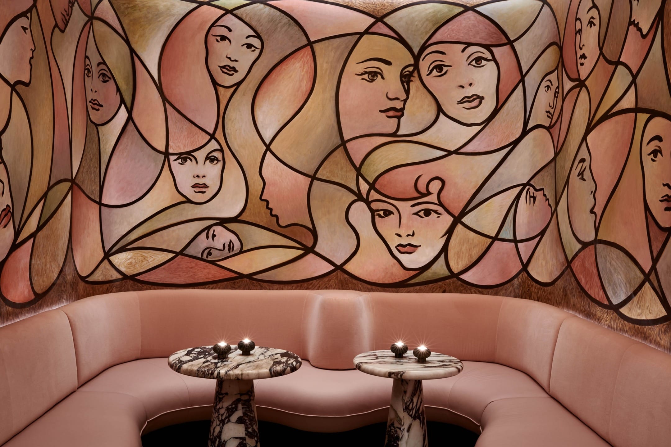 Abstract mural with faces in a cozy lounge setting featuring round marble tables and pink seating.