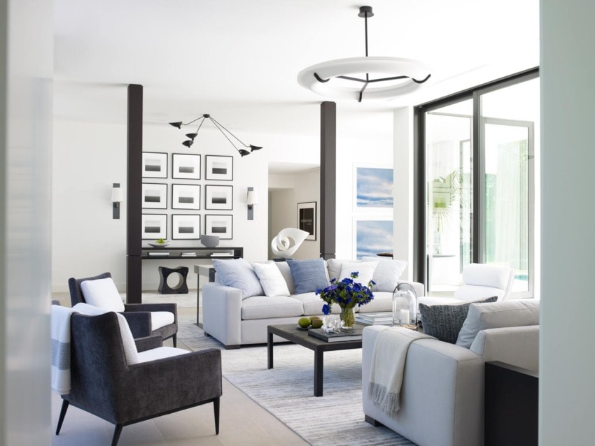 Modern living room with white sofas, black chairs, geometric lighting, and wall art, featuring large windows and a vase of flowers.